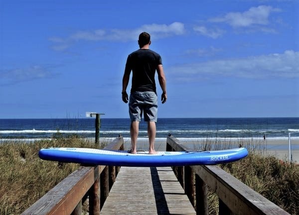 man standing on an inflatable paddle board that is laid across two sides of a deck path on the beach.